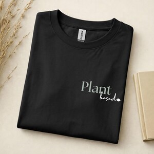 May include: A black crew neck t-shirt featuring the text "Plant" in a grey serif font and the word "based" in a white cursive script with a small leaf icon at the end. The shirt has a Gildan brand label visible at the inner neckline.