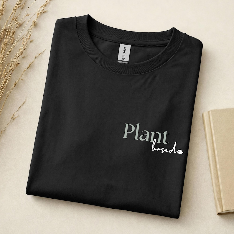 May include: A black crew neck t-shirt featuring the text "Plant" in a grey serif font and the word "based" in a white cursive script with a small leaf icon at the end. The shirt has a Gildan brand label visible at the inner neckline.