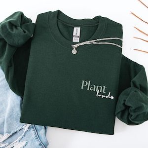 Könnte beinhalten: Ein dunkelgrünes Sweatshirt mit dem weißen Aufdruck "Plant based". Eine silberne Halskette mit Sonnenanhänger liegt über dem Sweatshirt. Das Sweatshirt liegt gefaltet auf einer Jeans.