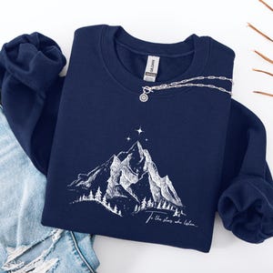 Könnte beinhalten: Marineblaues Sweatshirt mit weißer Berg-Grafik und dem Text "To the stars who listen...". Eine silberne Halskette mit Sonnenanhänger liegt auf dem Sweatshirt. Das Sweatshirt ist auf einer weißen Oberfläche gefaltet.