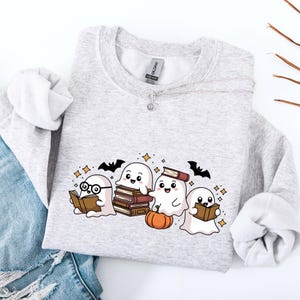 Könnte beinhalten: Hellgraues Sweatshirt mit einer Grafik von vier Cartoon-Geistern, die Bücher lesen, einem kleinen orangefarbenen Kürbis und Fledermäusen. Die Geister haben lächelnde Gesichter und sind von Sternen umgeben. Das Sweatshirt ist aus weichem Material.