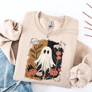 Könnte beinhalten: Beiges Sweatshirt mit einem Geistermotiv, umgeben von Blumen in einem braunen und schwarzen Rahmen. Der Geist hat ovale Augen und eine fließende Form. Die Blumen sind weiß, pfirsichfarben und rot. Eine silberne Halskette mit Sonnenanhänger befindet sich auf dem Sweatshirt.