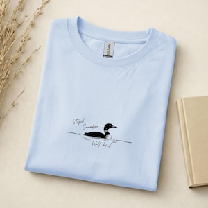 Pu&ograve; includere: Una t-shirt azzurro chiaro a girocollo con una grafica raffigurante una strolaga nera e bianca che nuota sull'acqua. Il testo "Stupid Canadian Wolf bird" &egrave; stampato in carattere corsivo nero vicino alla grafica. Etichetta del marchio Gildan.