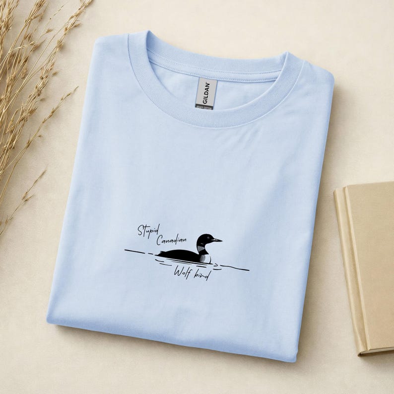 Pu&ograve; includere: Una t-shirt azzurro chiaro a girocollo con una grafica raffigurante una strolaga nera e bianca che nuota sull'acqua. Il testo "Stupid Canadian Wolf bird" &egrave; stampato in carattere corsivo nero vicino alla grafica. Etichetta del marchio Gildan.