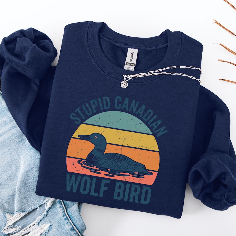 Puede incluir: Sudadera azul marino con un dise&ntilde;o retro que presenta un somorgujo en el agua y el texto "STUPID CANADIAN WOLF BIRD" en una fuente desgastada. El dise&ntilde;o incluye un gr&aacute;fico de puesta de sol colorido. Un collar plateado est&aacute; sobre la sudadera.