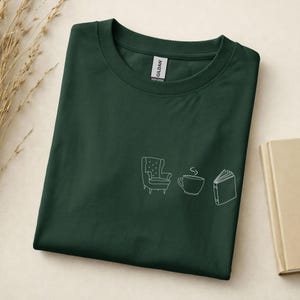 May include: A dark forest green crew neck t-shirt featuring a white line art graphic on the chest. The design consists of three simple icons arranged horizontally: an armchair, a steaming coffee cup, and an open book with visible pages.