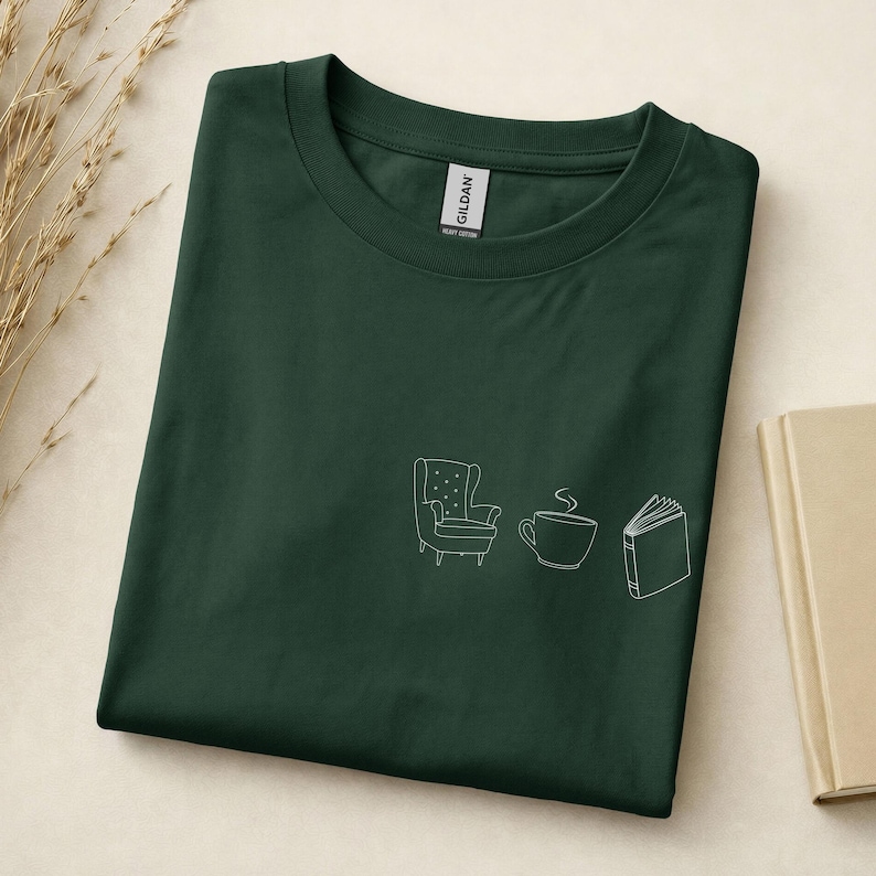 May include: A dark forest green crew neck t-shirt featuring a white line art graphic on the chest. The design consists of three simple icons arranged horizontally: an armchair, a steaming coffee cup, and an open book with visible pages.