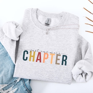 May include: A light gray heather crewneck sweatshirt featuring the text "just one more CHAPTER" printed in a multi-colored palette of dark teal, mustard yellow, terracotta, peach, and brown. The garment has a standard ribbed neckline and cuffs.