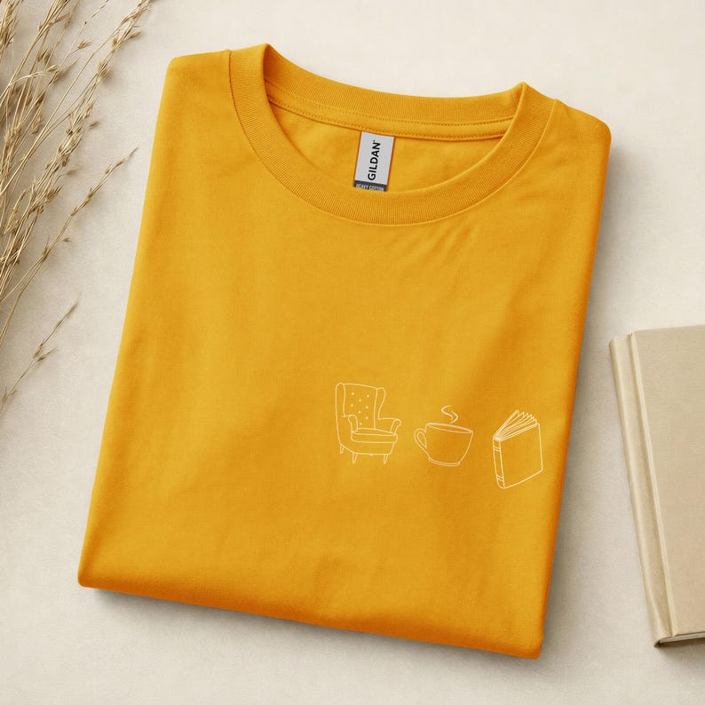 May include: A mustard yellow crew neck t-shirt featuring a small white line art graphic on the chest. The design depicts an armchair, a steaming cup of coffee, and an open book. The shirt has a Gildan brand label inside the collar.