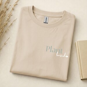 May include: A beige short-sleeve t-shirt with a crew neckline. The front features the text "Plant" in a muted green serif font and the word "based" in a white cursive script, accompanied by a small leaf icon next to the text.