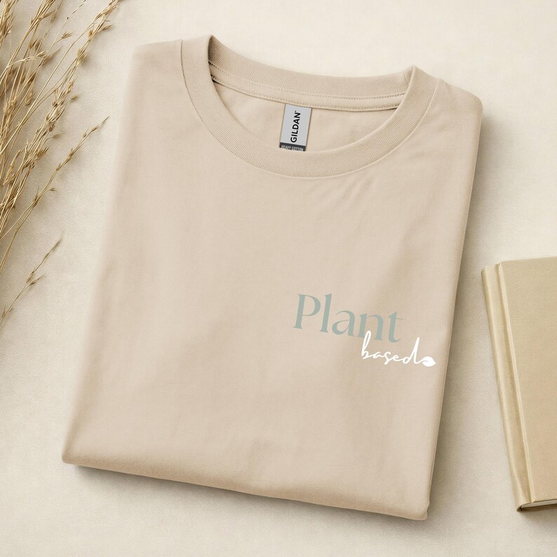 May include: A beige short-sleeve t-shirt with a crew neckline. The front features the text "Plant" in a muted green serif font and the word "based" in a white cursive script, accompanied by a small leaf icon next to the text.