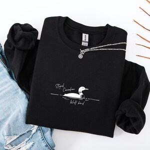Puede incluir: Sudadera negra con un gr&aacute;fico blanco de un somorgujo y el texto "Stupid Canadian Wolf bird". Un collar plateado con un colgante circular est&aacute; sobre la sudadera. La sudadera est&aacute; doblada sobre una superficie blanca.