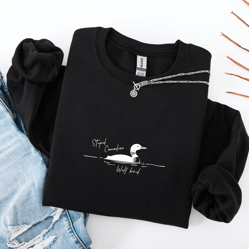 Puede incluir: Sudadera negra con un gr&aacute;fico blanco de un somorgujo y el texto "Stupid Canadian Wolf bird". Un collar plateado con un colgante circular est&aacute; sobre la sudadera. La sudadera est&aacute; doblada sobre una superficie blanca.