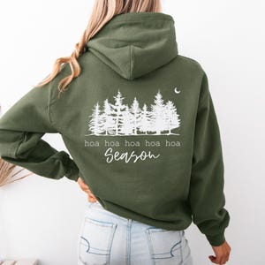May include: An olive green hooded sweatshirt featuring a white graphic on the back. The design displays a forest of pine trees, a small crescent moon, and the text "hoa hoa hoa hoa hoa Season" printed in a combination of serif and script fonts.