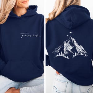 Könnte beinhalten: Marineblauer Kapuzenpullover mit Vordertasche. Auf der Vorderseite steht in weißer Schrift "To the stars who listen...". Auf der Rückseite befindet sich eine weiße Bergkette mit Bäumen und Sternen.
