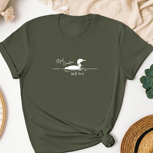 Pu&ograve; includere: Una t-shirt a maniche corte color verde oliva con una grafica bianca di un colimbo che nuota sull'acqua. Il testo "Stupid Canadian Wolf bird" &egrave; stampato in un carattere corsivo bianco sopra e sotto l'illustrazione dell'uccello sul petto.
