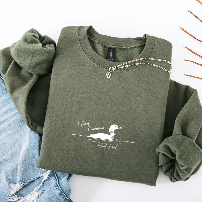 Puede incluir: Una sudadera verde oliva con el texto "Stupid Canadian Wolf bird" y un dibujo de un somorgujo. Un collar plateado est&aacute; colocado sobre la sudadera. La sudadera est&aacute; doblada junto a unos vaqueros azules.