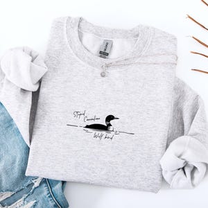 Puede incluir: Una sudadera gris claro con el texto "Stupid Canadian Wolf bird" y una ilustraci&oacute;n en blanco y negro de un colimbo. Un collar plateado con un peque&ntilde;o colgante est&aacute; sobre la sudadera.
