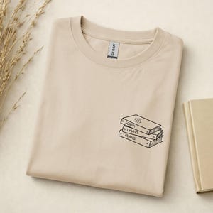 Könnte beinhalten: Ein hellbeiges T-Shirt mit Rundhalsausschnitt und einer kleinen schwarzen Strichzeichnung von drei gestapelten Büchern auf der Brust. Der Text auf den Buchrücken lautet SORRY I HAVE PLANS. Das Etikett der Marke Gildan ist am Halsausschnitt sichtbar.