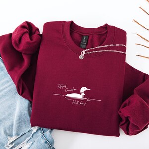 Puede incluir: Una sudadera granate con el texto "Stupid Canadian Wolf bird" y un dibujo de un somorgujo. Un collar plateado con un colgante circular est&aacute; sobre la sudadera. La sudadera est&aacute; doblada sobre unos vaqueros azules.