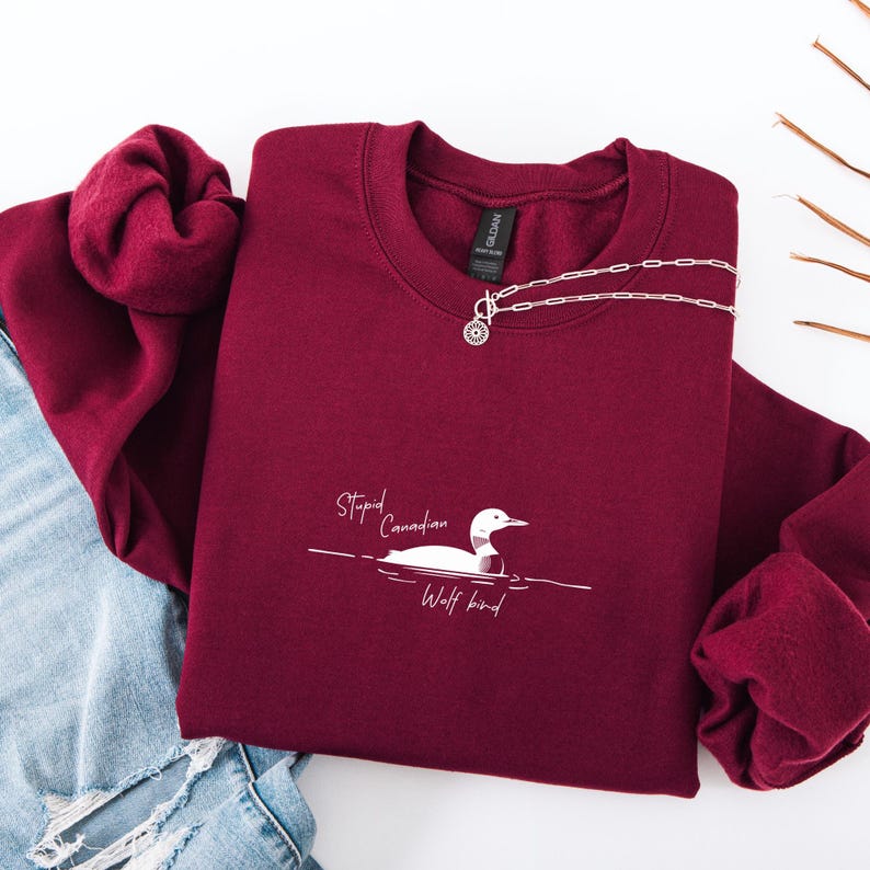 Puede incluir: Una sudadera granate con el texto "Stupid Canadian Wolf bird" y un dibujo de un somorgujo. Un collar plateado con un colgante circular est&aacute; sobre la sudadera. La sudadera est&aacute; doblada sobre unos vaqueros azules.