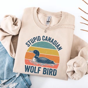 Puede incluir: Sudadera beige con el texto "STUPID CANADIAN WOLF BIRD" sobre un gr&aacute;fico de un somorgujo en un lago, sobre un dise&ntilde;o retro de puesta de sol. La sudadera est&aacute; doblada con un collar y unos vaqueros.
