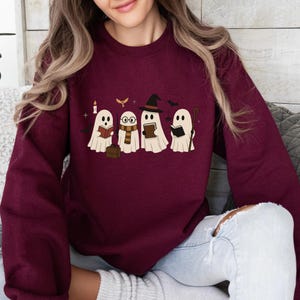May include: A maroon long-sleeve sweatshirt featuring a graphic print of four white ghosts reading books. The ghosts are depicted with various accessories including a witch hat, round glasses, a striped scarf, a wand, and a brown briefcase.