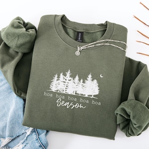 Könnte beinhalten: Ein olivgrünes Sweatshirt mit einer weißen Waldgrafik und den Worten "hoa hoa hoa hoa Season". Eine silberne Halskette mit einem Sonnenanhänger liegt auf dem Sweatshirt. Das Sweatshirt ist auf einer blauen Jeans gefaltet.
