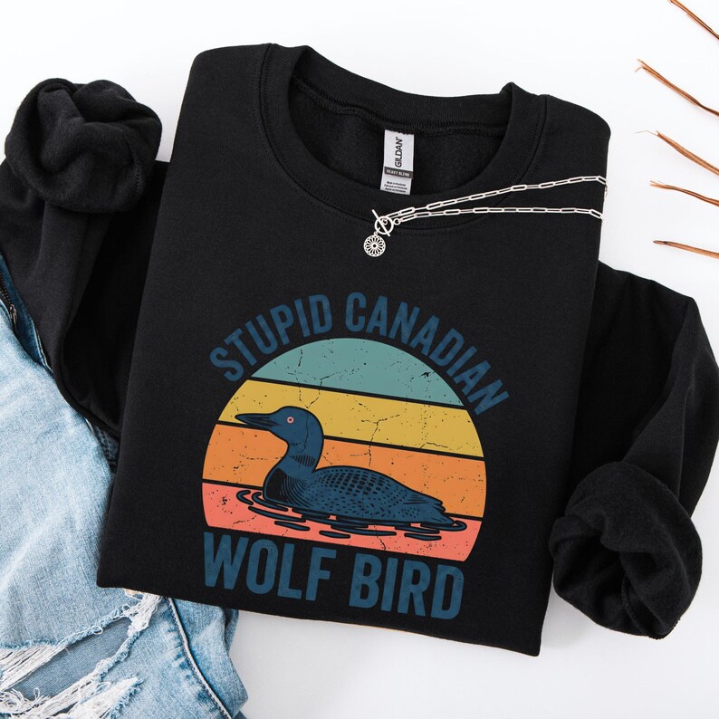 Puede incluir: Sudadera negra con un gr&aacute;fico de un colimbo en un dise&ntilde;o de puesta de sol. El texto dice "STUPID CANADIAN WOLF BIRD" en turquesa. Un collar plateado con un colgante circular est&aacute; drapeado sobre la sudadera. La sudadera est&aacute; doblada sobre unos vaqueros.