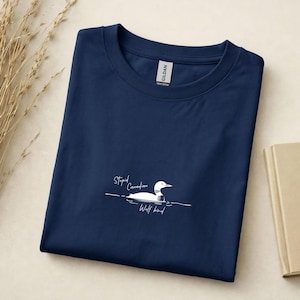 Pu&ograve; includere: Una t-shirt blu navy a girocollo con una grafica bianca di una strolaga che nuota sull'acqua. Il testo "Stupid Canadian Wolf bird" &egrave; stampato in un carattere corsivo bianco sotto l'uccello. La maglietta ha un'etichetta Gildan sul colletto.