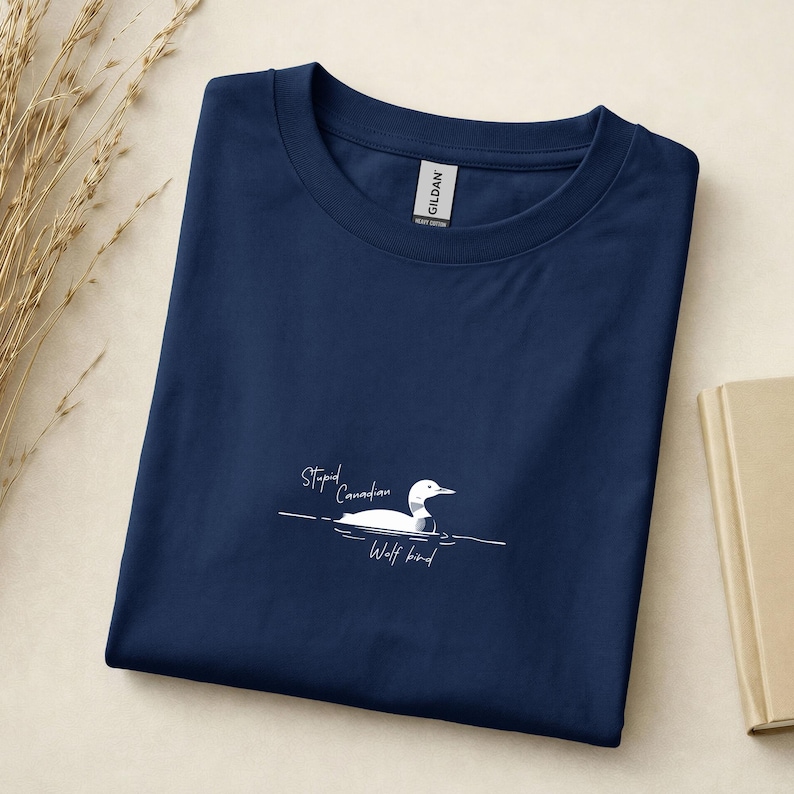 Pu&ograve; includere: Una t-shirt blu navy a girocollo con una grafica bianca di una strolaga che nuota sull'acqua. Il testo "Stupid Canadian Wolf bird" &egrave; stampato in un carattere corsivo bianco sotto l'uccello. La maglietta ha un'etichetta Gildan sul colletto.