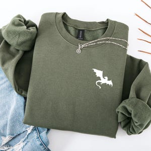 Op de afbeelding: Een olijfgroene sweatshirt met een witte drakenafbeelding op de voorkant. Een zilveren ketting met een cirkelvormige hanger is eroverheen gedrapeerd. De sweatshirt ligt op een spijkerbroek.
