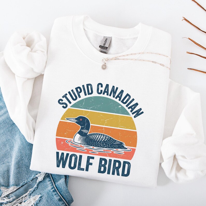 Puede incluir: Sudadera blanca con el texto "STUPID CANADIAN WOLF BIRD" en azul y blanco. El dise&ntilde;o presenta una ilustraci&oacute;n de somorgujo sobre un atardecer retro. Tambi&eacute;n se ve un collar plateado con un colgante.