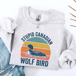 Puede incluir: Una sudadera gris claro con el texto "STUPID CANADIAN WOLF BIRD" en un dise&ntilde;o retro. El gr&aacute;fico presenta un colimbo grande frente a un fondo estilo puesta de sol con rayas turquesas, amarillas y naranjas. Un collar plateado est&aacute; drapeado sobre la sudadera.