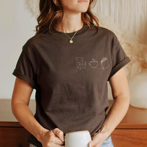 May include: A dark brown short-sleeved t-shirt featuring a small white line art graphic on the left chest. The design consists of three simple icons: an armchair, a steaming cup of coffee, and an open book, arranged in a horizontal row.