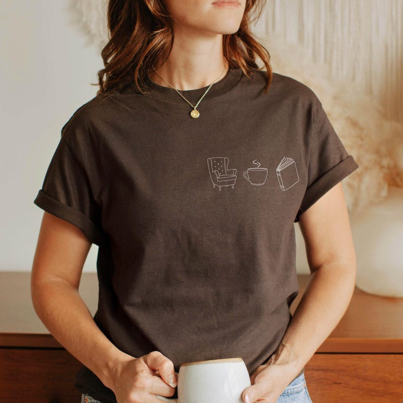 May include: A dark brown short-sleeved t-shirt featuring a small white line art graphic on the left chest. The design consists of three simple icons: an armchair, a steaming cup of coffee, and an open book, arranged in a horizontal row.
