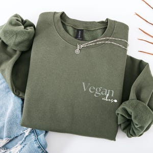 Op de afbeelding: Een olijfgroene sweatshirt met de woorden "Vegan vibes" in lichtgrijze letters. Een zilveren ketting met een kleine hanger is over de voorkant gedrapeerd. De sweatshirt is op een spijkerbroek gevouwen.