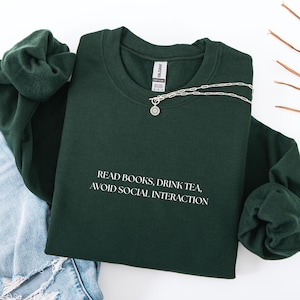 Könnte beinhalten: Ein dunkelgrünes Sweatshirt mit dem weißen Text "READ BOOKS, DRINK TEA, AVOID SOCIAL INTERACTION". Eine silberne Halskette mit Sonnenanhänger liegt über dem Sweatshirt. Das Sweatshirt liegt auf einer Jeans.