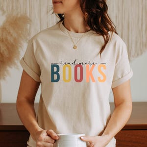Könnte beinhalten: Beiges T-Shirt mit dem Schriftzug "read more BOOKS" in einer farbenfrohen, verspielten Schriftart. Das Wort "read more" steht in einer Schreibschrift über dem Wort "BOOKS". Das Shirt hat hochgekrempelte Ärmel und einen Rundhalsausschnitt.