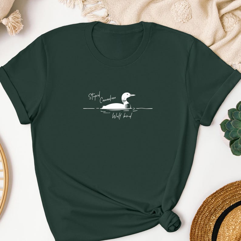 Pu&ograve; includere: Una t-shirt a maniche corte color verde bosco scuro con una grafica bianca di una strolaga che nuota sull'acqua. Il testo "Stupid Canadian Wolf Bird" &egrave; stampato in un carattere corsivo bianco sopra e sotto l'illustrazione dell'uccello.