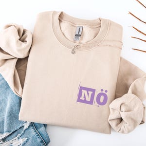 Op de afbeelding: Een beige sweatshirt met het woord "NÖ" in paars, een zilveren ketting en een spijkerbroek. De sweatshirt is op een wit oppervlak gevouwen. De tekst staat in een paarse rechthoek.