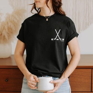 Könnte beinhalten: Ein schwarzes Kurzarm-T-Shirt mit einer weißen Grafik im linken Brustbereich. Die Grafik zeigt zwei gekreuzte Hockeyschläger, einen Puck und die Zahlen 22 44 66 46, die in einer klaren, serifenlosen Schrift unter den Schlägern gedruckt sind.