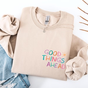 Könnte beinhalten: Beiges Sweatshirt mit Rundhalsausschnitt und dem Schriftzug "GOOD THINGS AHEAD" in bunten Farben. Eine silberne Halskette mit Sonnenanhänger liegt auf dem Sweatshirt. Das Sweatshirt ist mit einer Jeans und einem beigen Ärmel gefaltet.