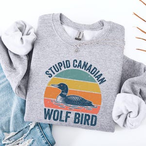 Puede incluir: Una sudadera gris con el texto "STUPID CANADIAN WOLF BIRD" sobre un gr&aacute;fico de un somorgujo en un dise&ntilde;o retro de puesta de sol. La sudadera est&aacute; doblada con unos vaqueros azules.