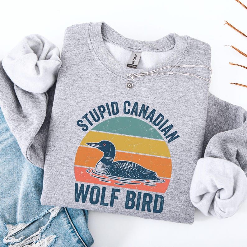 Puede incluir: Una sudadera gris con el texto "STUPID CANADIAN WOLF BIRD" sobre un gr&aacute;fico de un somorgujo en un dise&ntilde;o retro de puesta de sol. La sudadera est&aacute; doblada con unos vaqueros azules.