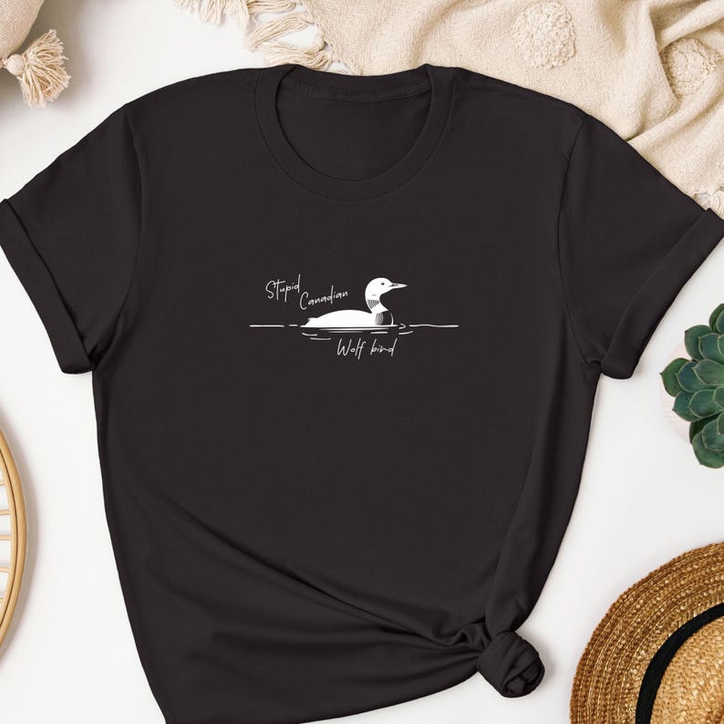 Pu&ograve; includere: Una t-shirt nera a maniche corte con una grafica bianca raffigurante un colimbo che nuota sull'acqua. Il testo "Stupid Canadian" &egrave; stampato sopra l'uccello, mentre le parole "Wolf bird" sono scritte sotto in un carattere corsivo bianco.