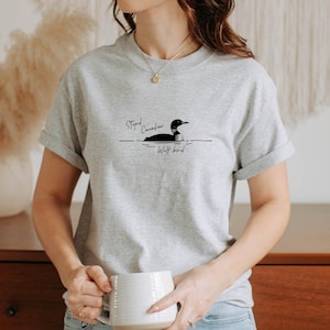Pu&ograve; includere: Una t-shirt grigio chiaro a maniche corte con una grafica nera di un colimbo che nuota sull'acqua. Il testo sopra l'uccello recita "Stupid Canadian" e il testo sottostante "Wolf bird" in un carattere corsivo nero. Scollo rotondo.