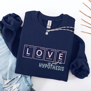 Könnte beinhalten: Ein marineblaues Sweatshirt mit Rundhalsausschnitt und einem Grafikdruck des Wortes LOVE, dargestellt als Elemente des Periodensystems in hellrosa Kästchen mit weißer Schrift. Darunter steht "is just a HYPOTHESIS" in grauer Schrift.