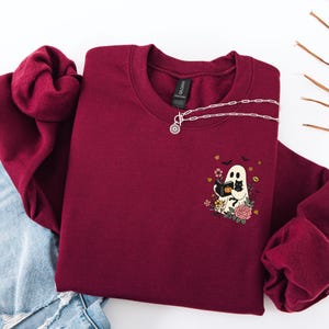 Könnte beinhalten: Burgunderfarbenes Sweatshirt mit Rundhalsausschnitt und einem Aufdruck eines Geistes, der ein Buch liest, einer schwarzen Katze, Blumen und Fledermäusen. Eine silberne Halskette mit einem Sonnenanhänger liegt auf dem Sweatshirt. Das Sweatshirt liegt auf einer weißen Oberfläche.