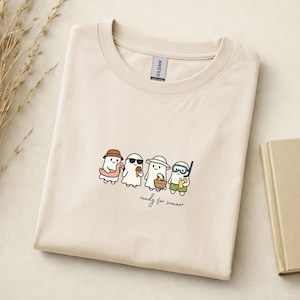 May include: A light beige t-shirt featuring a graphic of four cartoon ghosts dressed for summer activities. The ghosts wear a bucket hat with a pink floatie, sunglasses with an ice cream cone, a sun hat with a beach bag, and snorkel gear with a drink.